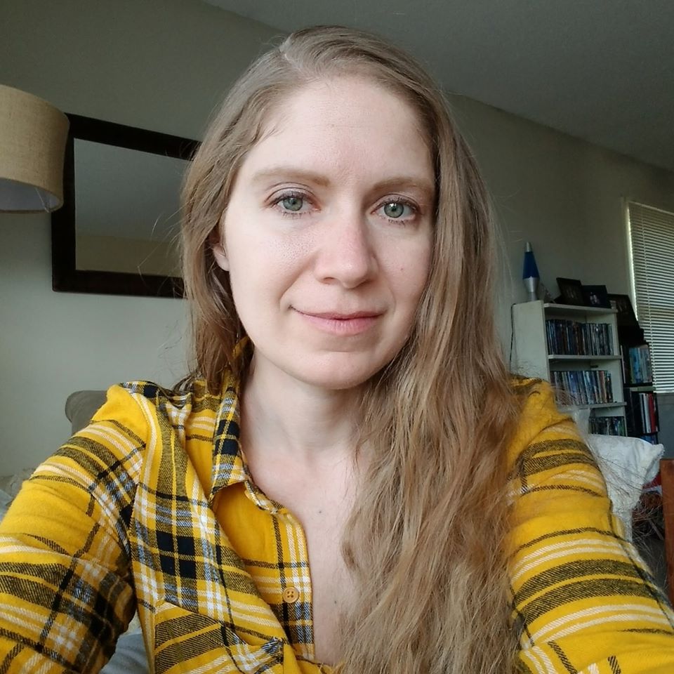 Photo is of Kelly wearing a yellow and black plaid shirt. She has long blond hair parted on the side.