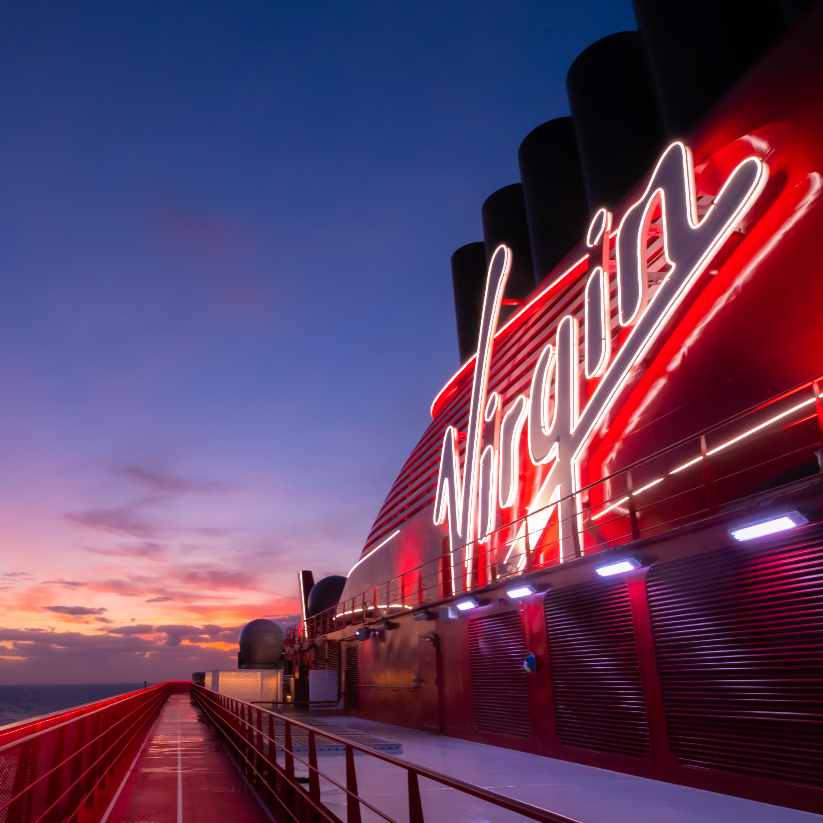 Image shows a photo of the Virgin Voyages cruise ship, Scarlet Lady, at sunset. The logo is brightly lit.