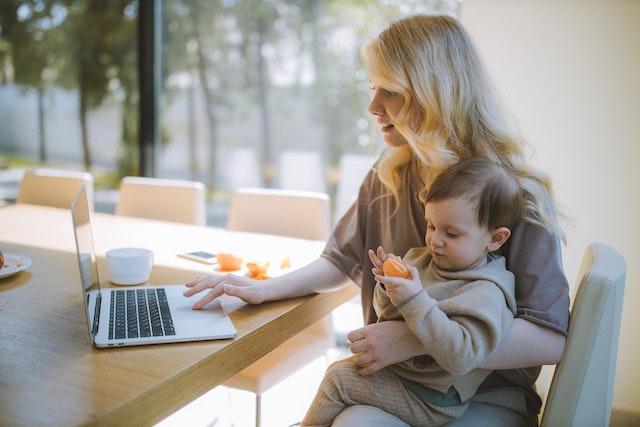 A mom works from her kitchen table on a laptop while holdig her child. Photo by Anastasia Shuraeva.