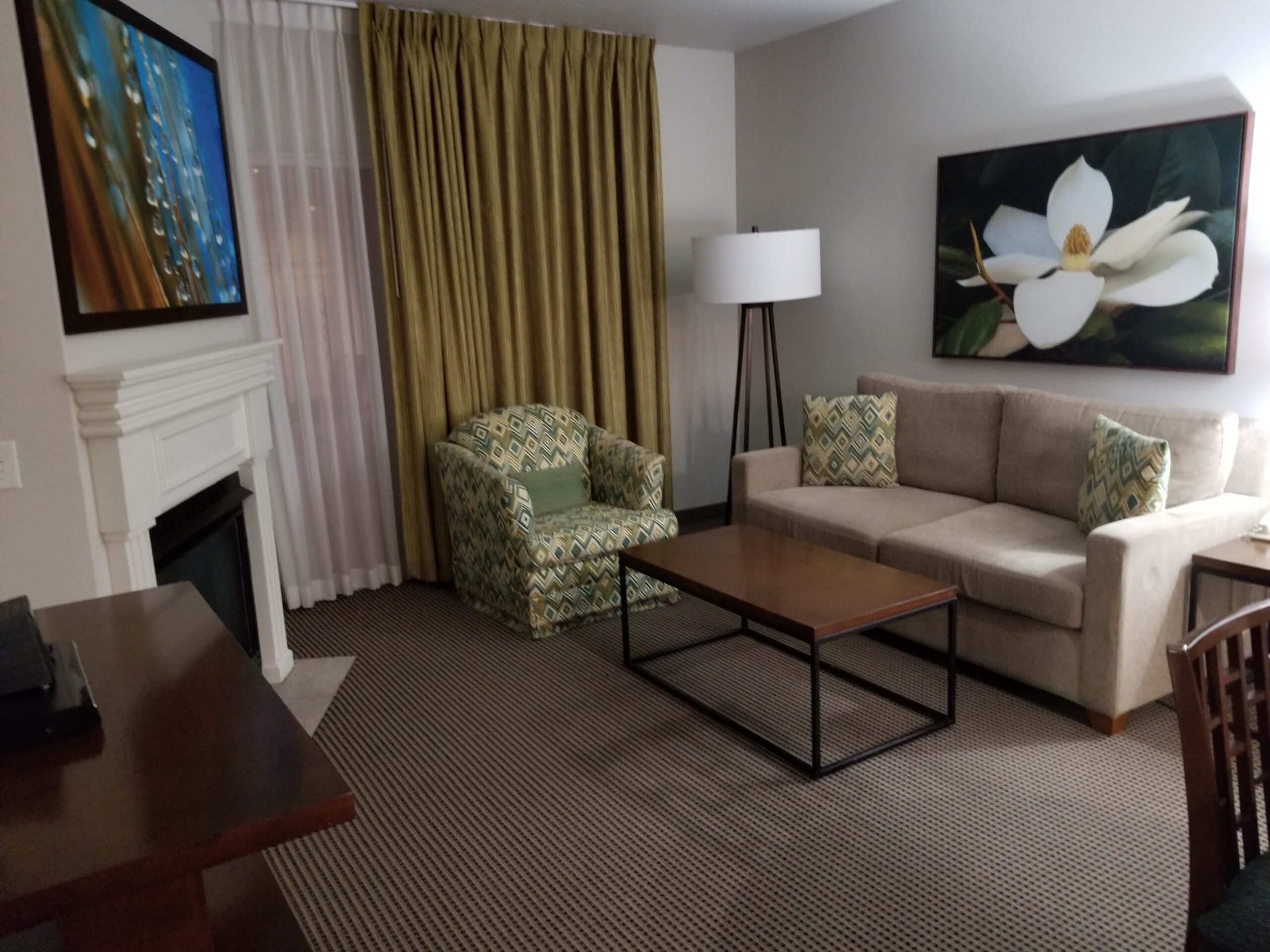 This photo features the living room area in a suite at the Tahiti resort on Tropicana Boulevard in Las Vegas. It has light brown carpet, a dark brown wooden desk on the left wall, a cream colored couch and painting of a white flower on the left wall, a coffee table with a wooden top and a black metal frame underneath, a cushioned chair against the gold curtain on the back wall. There is a white curtain stretched all the way across the window and behind the gold curtain which separates the living room from the balcony. there is also a gas fireplace with a white mantel on the left wall and a painting above that.