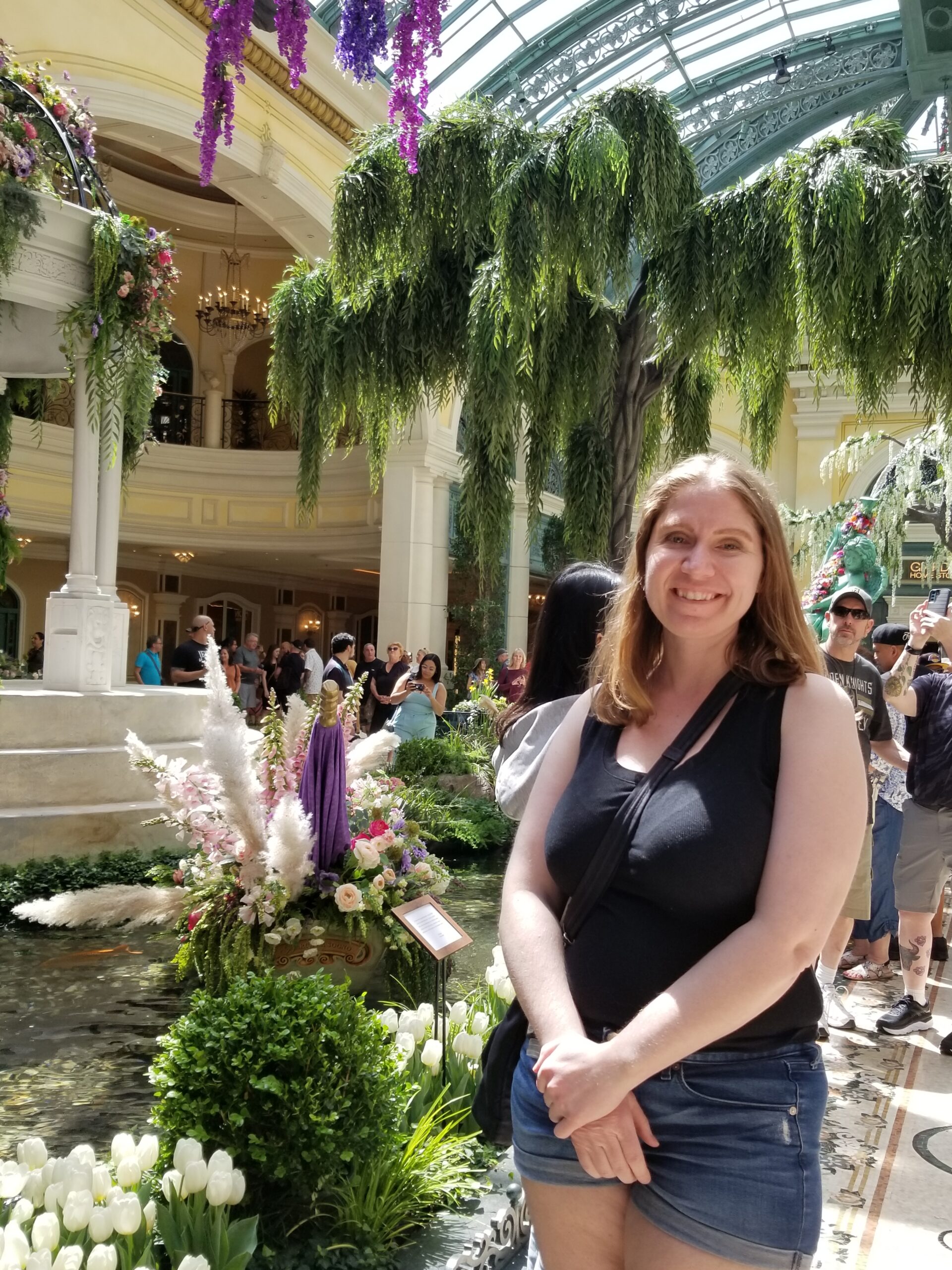 The author, an early forties, but much younger-looking, caucasian female with medium-length strawberry blonde hair clasps her hands while standing at a slight angle to the camera and smiling. She is in the garden at the Bellagio hotel in Las Vegas with sunlight coming in from above. She is wearing a black tank top and denim shorts and is carrying a black purse. 