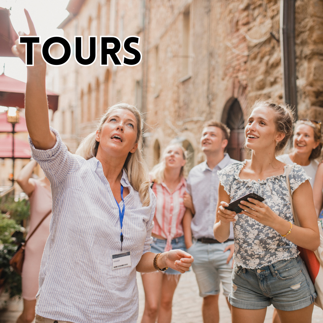 This image features a tour guide pointing up and to the viewer's left to show her guests something. The group, made up of four visible guests, who are all caucasian adults, are all outside and an old tan-colored building are seen behind them. The word "Tours" is superimposed in the top left corner in black text in all caps.